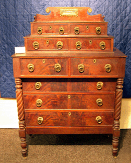 A chest of drawers in mahogany veneer over pine had an interesting double stepped back bank of drawers with a fancifully wrought brass plate in the center of the backsplash as seen at Carlson and Stevenson, Manchester, Vt.