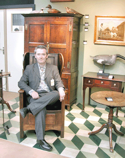 Martin Murray, London, who specializes in period country furniture and folk art, sits in his booth on an early Nineteenth Century lambing chair from Lancashire. Farmers would use the chair to nurse new-born lambs who needed extra attention, with the drawer beneath the seat serving as a warm, protective "bed†for the infant animal.
