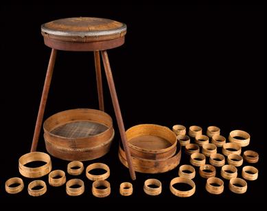 A group of sieves, produced at Mount Lebanon in the early to mid-Nineteenth Century, is on view with a sieve binding frame on a three-legged platform. The binding frame was used to hold the woven mat between the wooden hoops that held the sieve together. A collection of sieves, most of which are unfinished, surround the stand, illustrating the variety of sizes available.