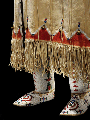 A detailed view of the bottom of a Southern Cheyenne beaded dress, circa 1930, shows the leggings and moccasins. The dress and accessories are made of hide, glass seed beads, mescal beans, brass bells, tin cones, fire-polished beads and red paint. They are probably from Oklahoma.