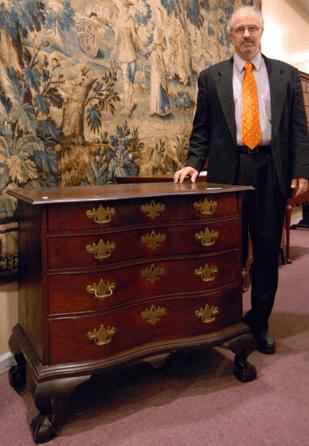 John Fontaine stands with the top lot of the sale, a reverse serpentine mahogany Chippendale four-drawer ball and claw foot chest that sold for $34,500. In the background, a 171-by-109-inch handmade scenic tapestry, Nineteenth Century, brought $6,038.