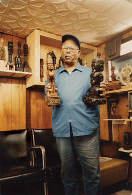 A pipe-smoking Ulysses Davis proudly displays two of his wood sculptures, with others in the background of his barbershop, where he also did his carving. Courtesy High Museum of Art.