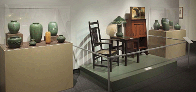 A view of the exhibition area showing the integration of furniture with accessories from the Arts and Crafts period. Furniture includes a Harvey Ellis-designed inlaid chair and fall front desk by Gustav Stickley and a Grueby tile-top table. Grueby pottery fills the cases on either side.