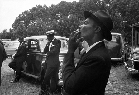 In "Funeral †St Helena, South Carolina,†1955, three well-dressed African American men waiting outside a rural funeral in the South look off to the left, observing events outside the frame. Their dignified and attentive appearance belies their status as second-class citizens. Susan and Peter MacGill. ⁐hotograph ©Robert Frank, from The Americans