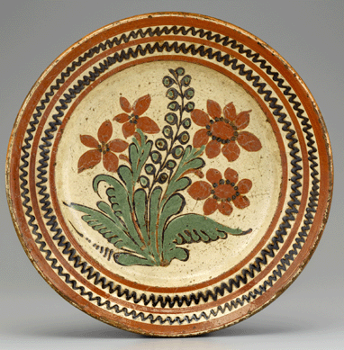 Many of the dishes associated with the tenure of Gottfried Aust (1722‱788) as master of the Salem pottery have centers with naturalistic flowers trailed in two, three or four colors of slip. As the leaves and flowers on this dish of 1775 to 1785 reveal, Aust and his workmen were highly skilled in their application of slip. The floral motif may have been inspired by a botanical illustration of the time. Lead-glazed earthenware, diameter 13¾ inches. Old Salem Museums & Gardens.