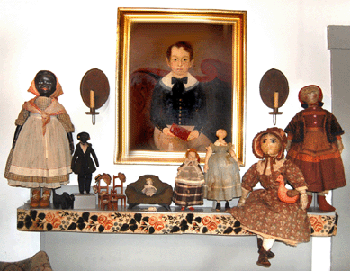 Rare black dolls, with miniature furniture and other cloth dolls, perch on a shelf beneath a folk portrait of a boy.