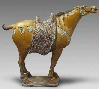 The noble horse, symbol of wealth and power, is adorned with saddle and decorative tack. From the Tang dynasty, it is of cream earthenware with a three-color glaze. Gift of Sadajiro Yamanaka.