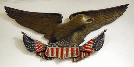 A finely carved and paint decorated spread-wing eagle holding a pair of flags and a shield.