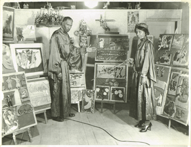 In this circa 1940 photograph, Elijah Pierce and his wife Cornelia, dressed in ministerial robes, pose in front of a selection of the artist's carved and painted lessons and bible stories. A tableau version of "Crucifixion†is mounted on the wall above Pierce's right shoulder. He used his carved objects to illuminate points when he preached. Columbus Museum of Art, Elijah Pierce Archive.