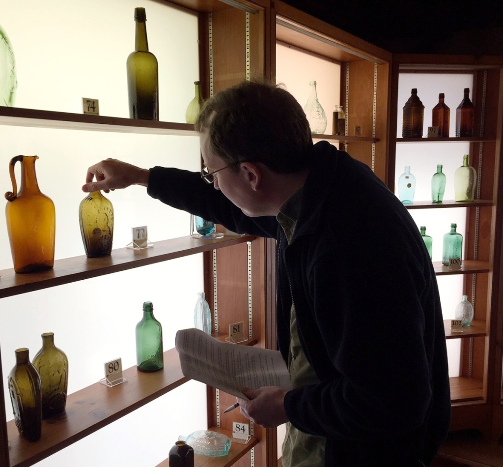 A buyer checks out a “Lafayette” and bust/liberty cap pint portrait flask in olive amber, $1,112. Above, a “Willington Glass Works” quart whiskey bottle in medium olive amber, $293, and, below, a “Saratoga / A / Spring Co. / N.Y.” pint mineral-water bottle in yellow green, $117. Bottom shelf, right, is a “General Lafayette” and bust with eagle and “T.W.D.” portrait flask in aquamarine, $234.