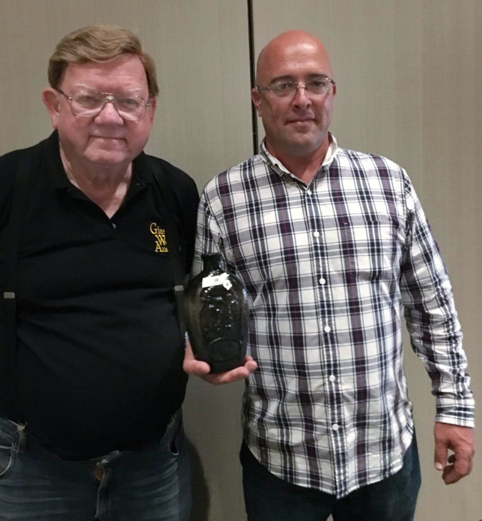 Jim Hagenbuch (left) and Jesse Sailer show off the auction’s top-selling lot, an eagle Masonic flask in a pale, almost clear olive amber color, Keene (N.H.) Glass Works, that sold for $9,360 to a floor bidder.