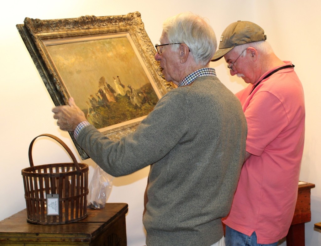 Dana Tillou, left, and Dave Brown try (unsuccessfully) to decipher the artist’s signature on this painting of seaside strollers in the booth of Maggie Milgrim Fine Art & Antiques, New York City.