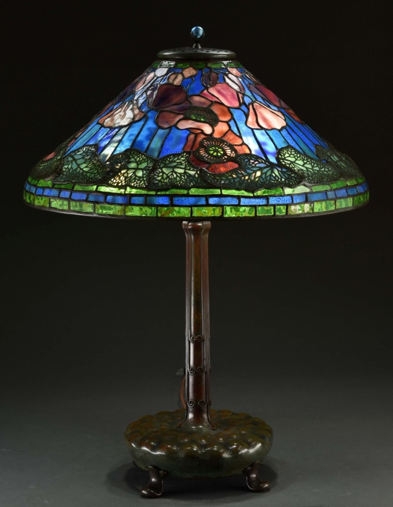 A Tiffany Studios Poppy table lamp measures 27 inches tall with a shade that is 20¼ inches in diameter. This lamp has a leaded glass shade in purple and maroon poppies set against a mottled and shaded blue background. A band of shaded green poppy leaves surround the edge of this shade. This lamp sold within estimate at $141,450.