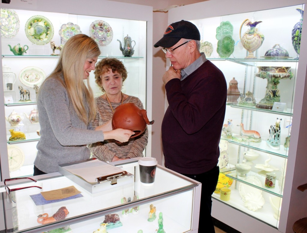 Customers examine an Eighteenth Century English ceramic redware pottery piece at at the booth of Monroe, Conn., dealer Maria and Peter Warren Antiques, while proprietor AJ Warren (center) answers their questions.