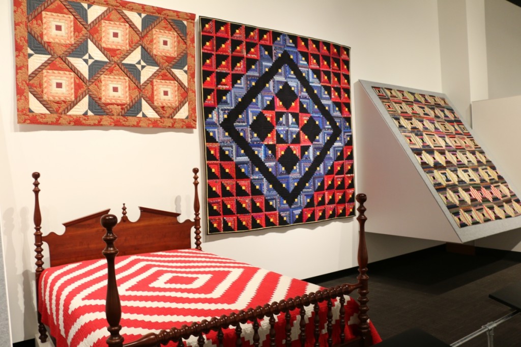 Two versions of a Log Cabin pattern quilt are displayed side by side. On the bed, a Barn Raising variation made in the 1920s in Rutherford County. Cotton with cotton batting; hand pieced. On the wall, a late-Twentieth Century Star variation by Charles Goddard (1927–2012) made in Memphis, Shelby County. Cotton with polyester batting; hand pieced.