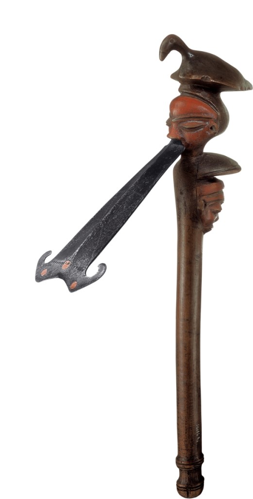 This ceremonial adze, with its tonguelike iron blade, once dignified the left shoulder of a Pende chief who wore it as an emblem of high office when traveling. Never intended to carve wood, the adze represented smooth-cutting diplomacy, straight talk, and efficient negotiation. The bird at the top alludes to the chief’s bird’s-eye view, or oversight, of his domain. In contrast, the calm, masklike face from which the blade emerges is protected by another set of eyes, keeping watch behind him. Adze by Central Pende artist, Democratic Republic of the Congo, early Twentieth Century. Iron, wood, copper, pigment. Felix Collection.