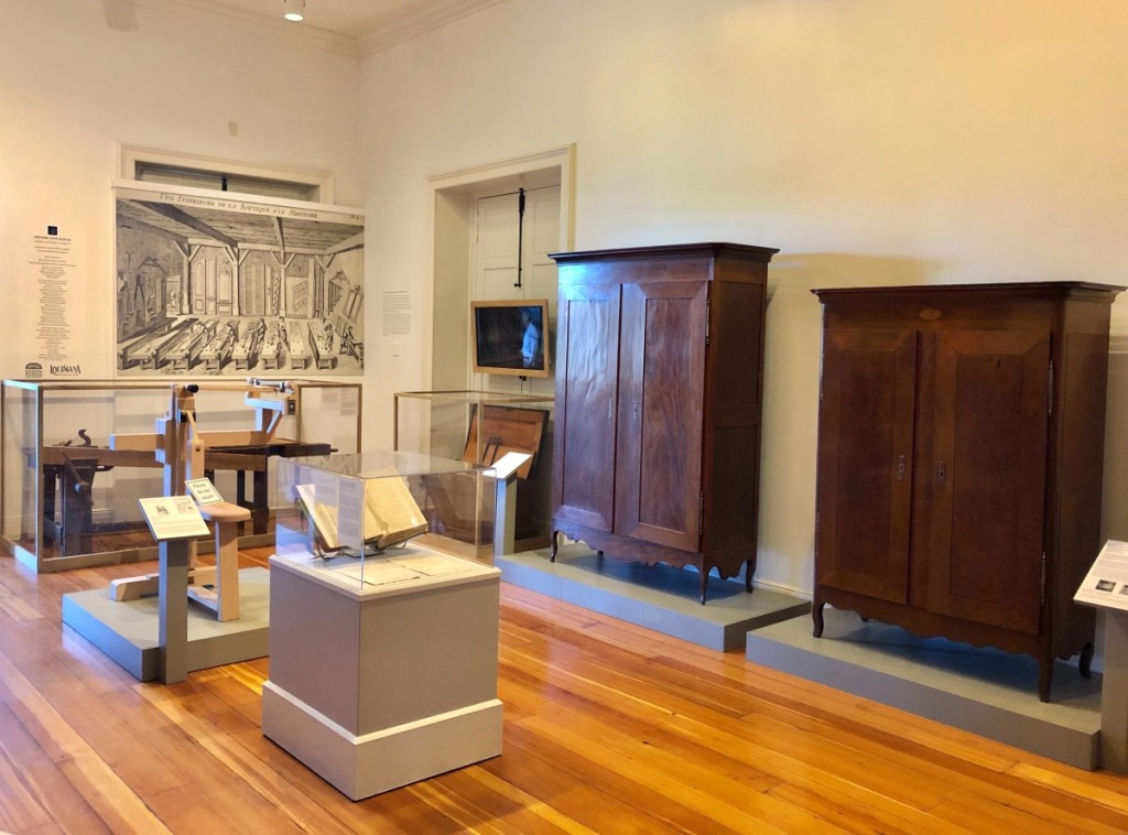 An installation view shows two Butterfly Man armoires along with objects that demonstrate their context. The left back wall displays a reproduction of “Vue Interieure de la Boutique d’un Menuisier,” an engraved image that appears in André Jacob Roubo’s  L’Art du Menuisier, featuring elements of a cabinetmaker’s workshop in the late Eighteenth Century. Beneath it is a Nineteenth Century cabinetmakers bench along with period tools.