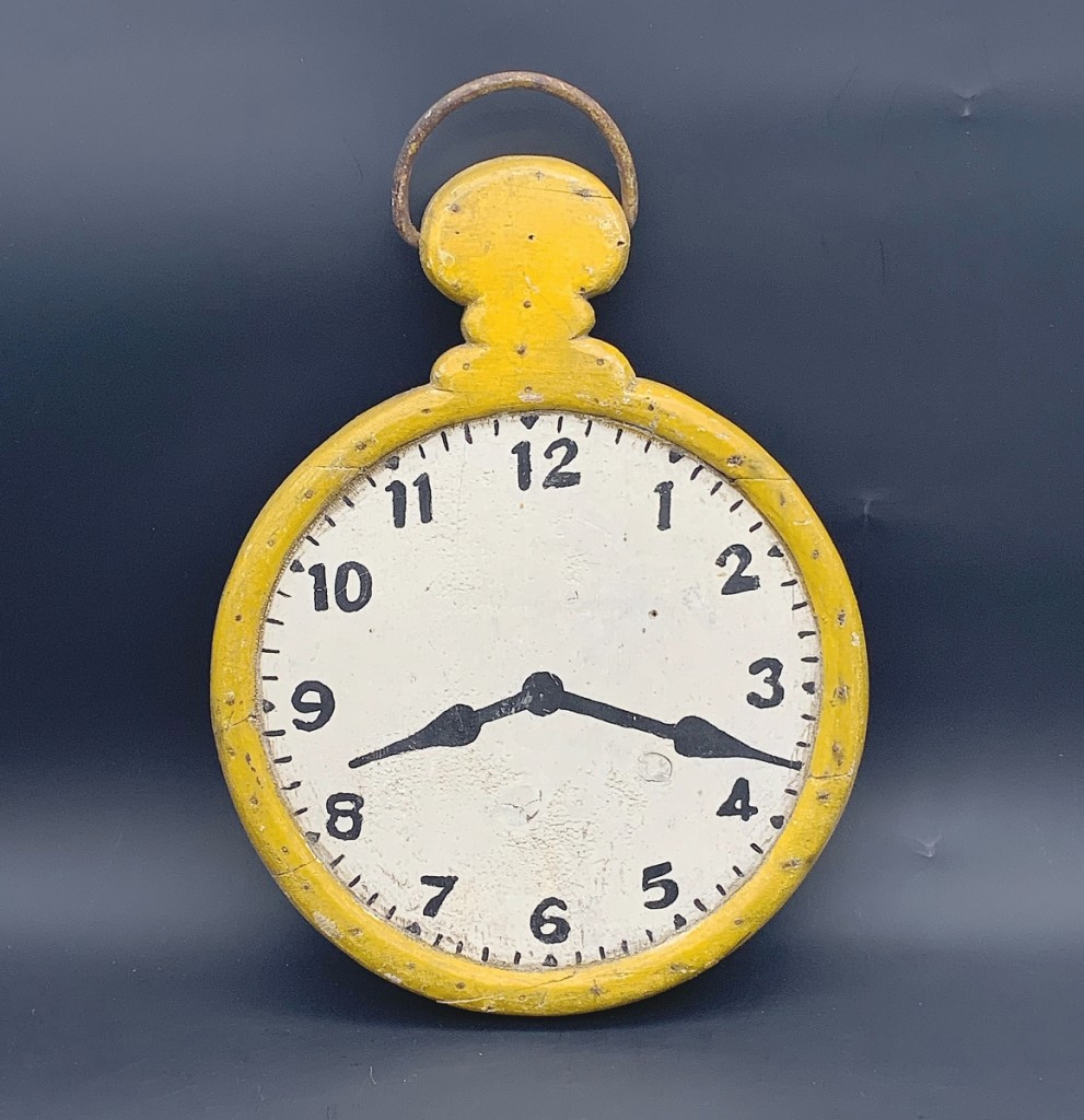 A wooden pocket watch trade sign from the early Twentieth Century with yellow painted wooden case and white and black painted dial connected with a buyer. Measuring 16 by 11 inches and in untouched original condition, it was offered by Mary & Joshua Steenburgh.