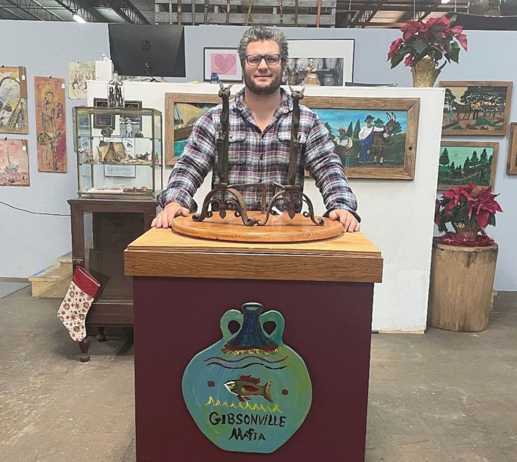 Auctioneer Matt Ledbetter stands at his podium with a pair of rabbit-form andirons that went out at $1,353. He said they were the best pieces of folk art in the sale. To the front of the podium is a placcard for the Gibsonville Folk Art Mafia, a gang of folk art collectors and sellers, which includes Matt Ledbetter, his wife and kids, his father Wade, Glen Richardson and Ron Causey Jr.