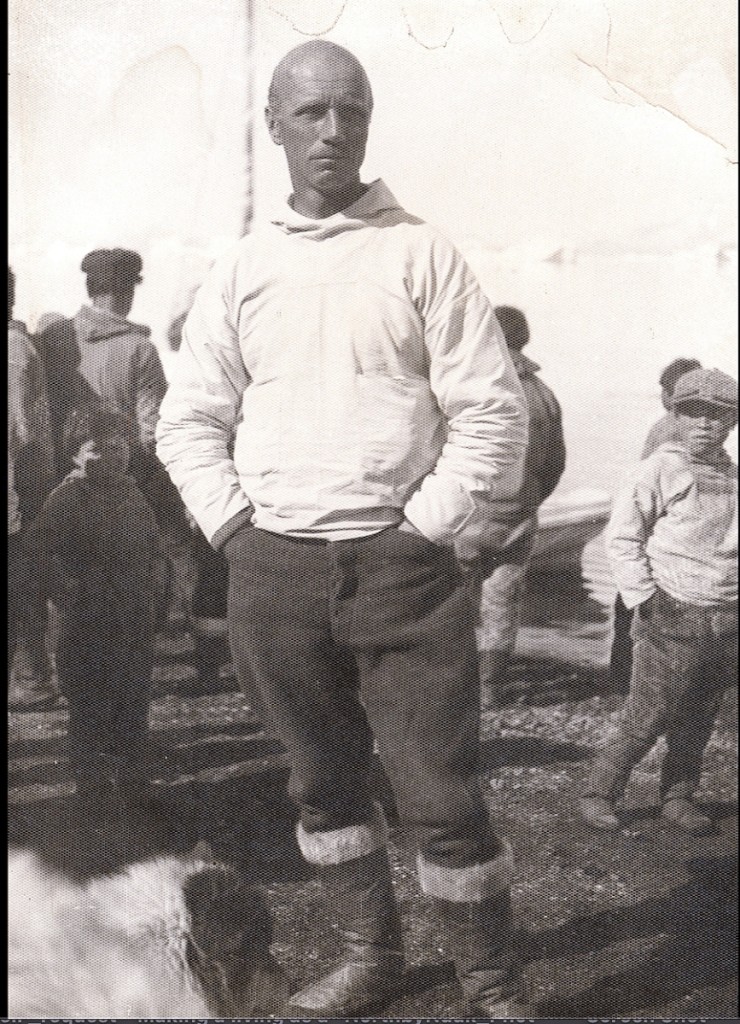 Photo of Rockwell Kent wearing anorak and seal skin boots, possibly on the Illorsuit beach with Inuit residence, circa 1932.   —Kent photos